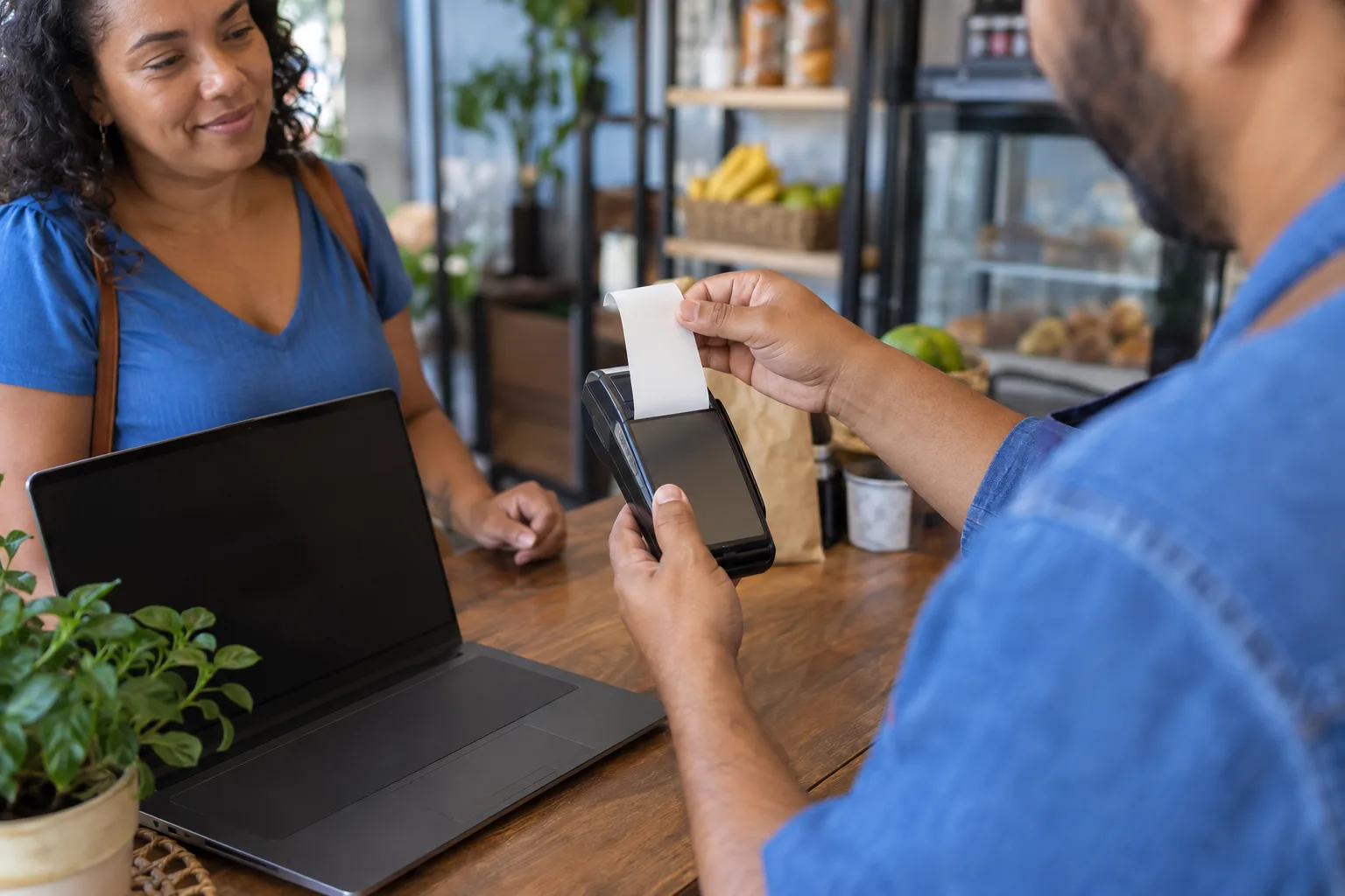 Mãos seguram o comprovante da maquininha ao lado do notebook no balcão