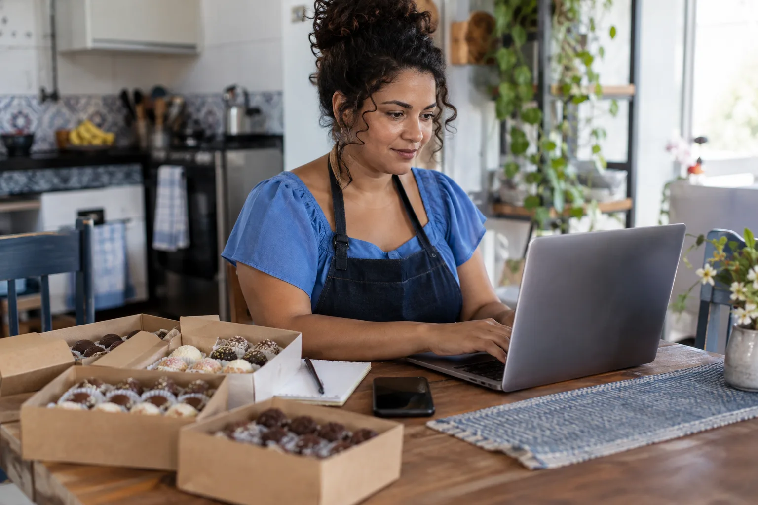 Doceira MEI trabalha no notebook na mesa da cozinha ao lado de caixas de doces