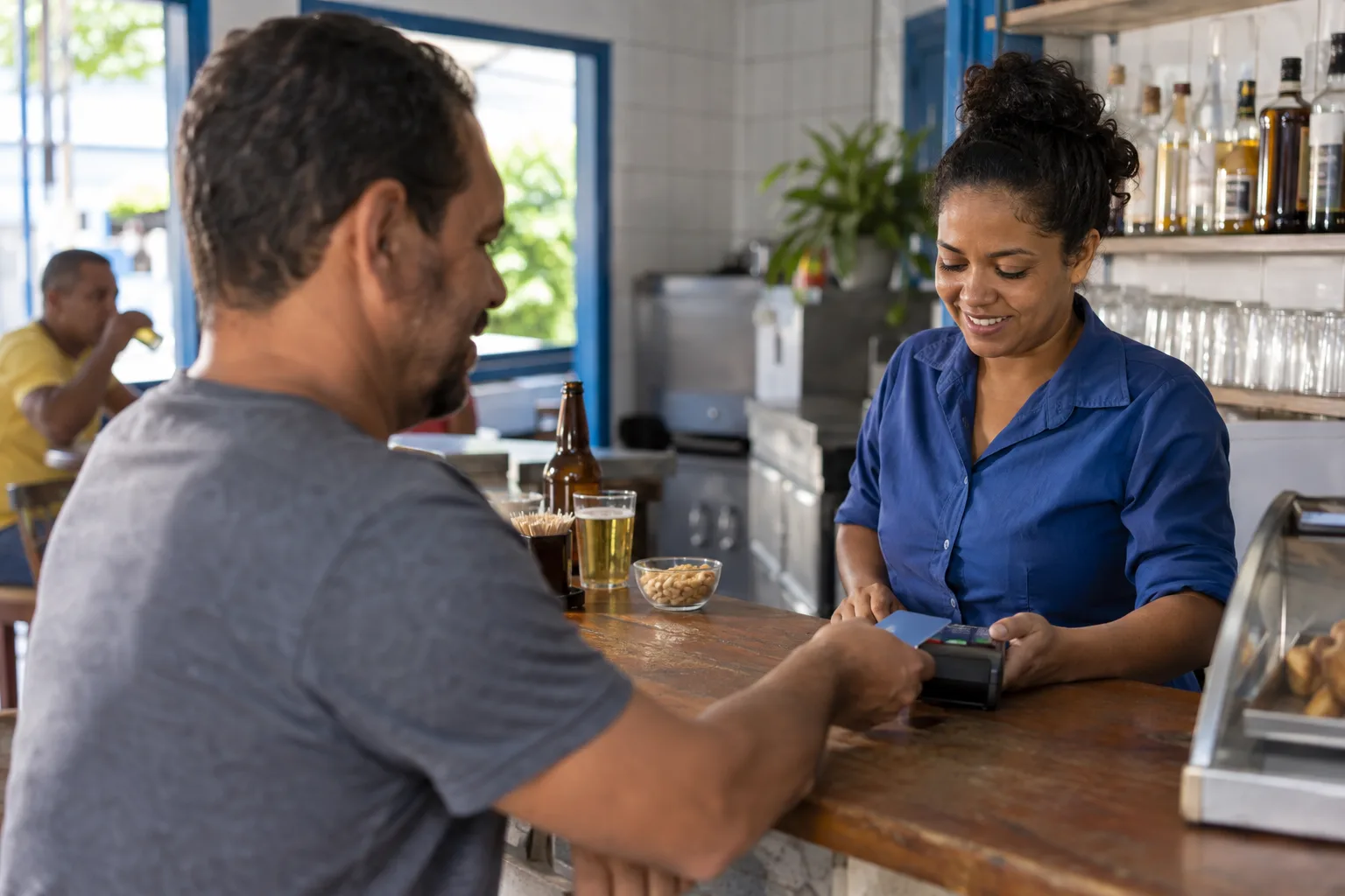 Atendente de bar fecha a conta de um cliente no balcão com a maquininha de cartão