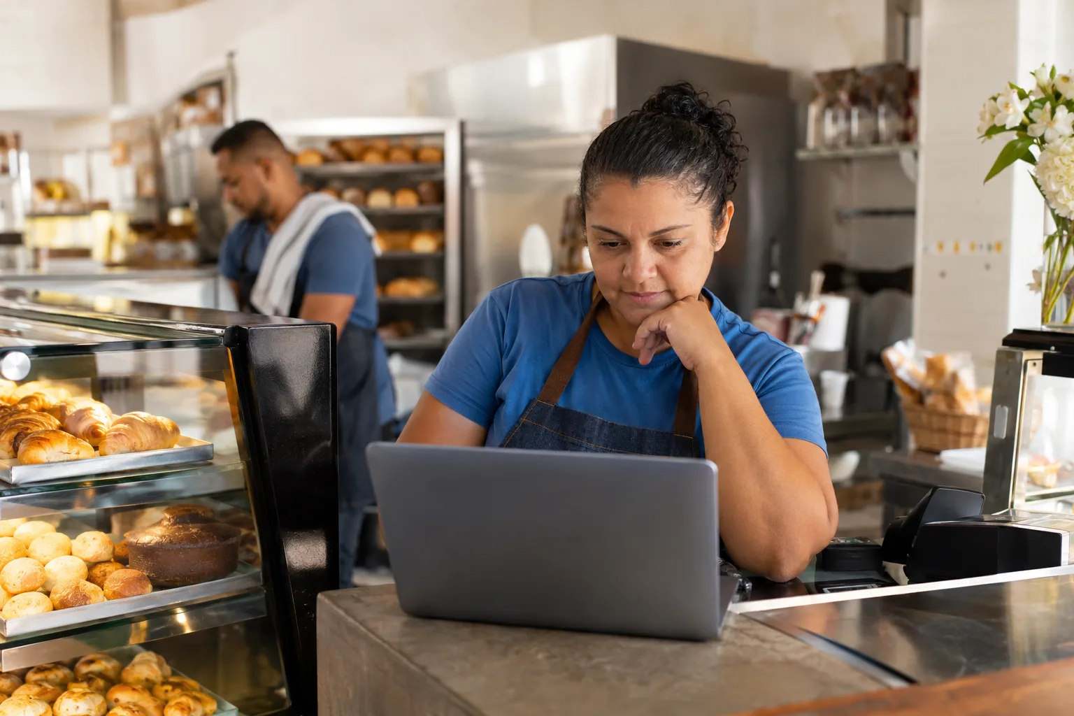 Dona de padaria consulta o portal de impostos no notebook ao lado do caixa