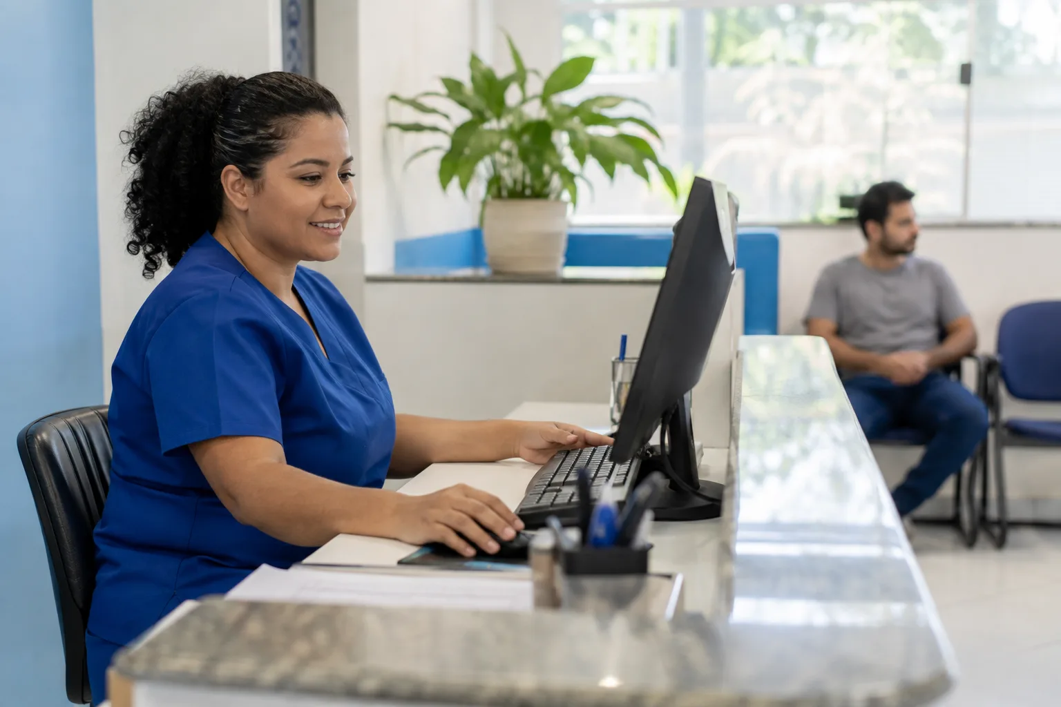 Recepcionista de clínica trabalha no computador do balcão enquanto um paciente aguarda